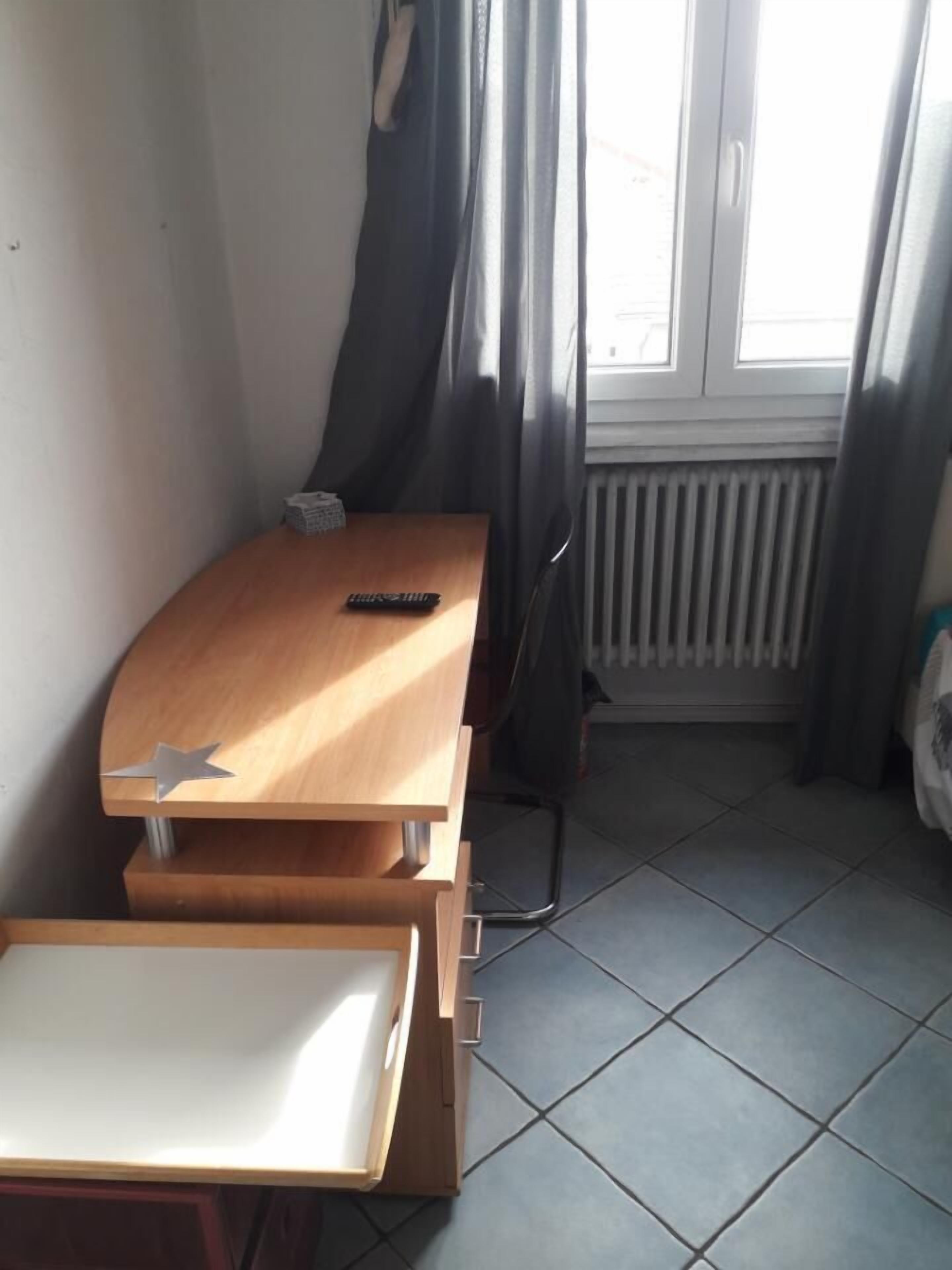 Habitación pequeña con escritorio de madera, una silla, radiador y ventana con cortinas.
