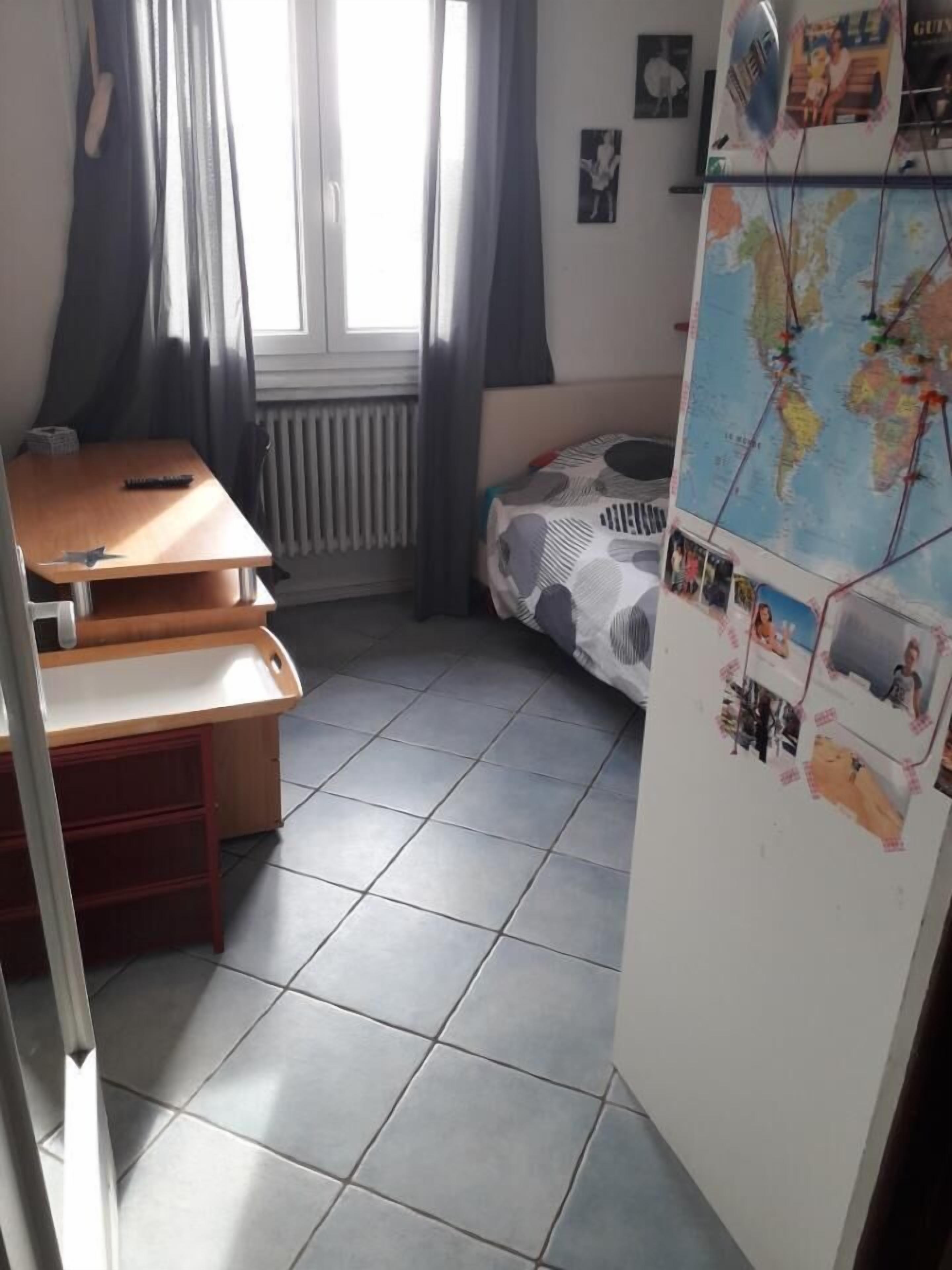 Una habitación con cama, escritorio, silla, radiador, ventana con cortinas y una pared con un mapa del mundo y fotografías.