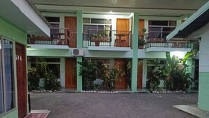 A two-story building with a sign reading "HOTEL" above the entrance. There are multiple balconies with potted plants, and the ground floor has a gravel courtyard. - Hotel FAS & OFF Site Thermal Resort (La Fortuna)