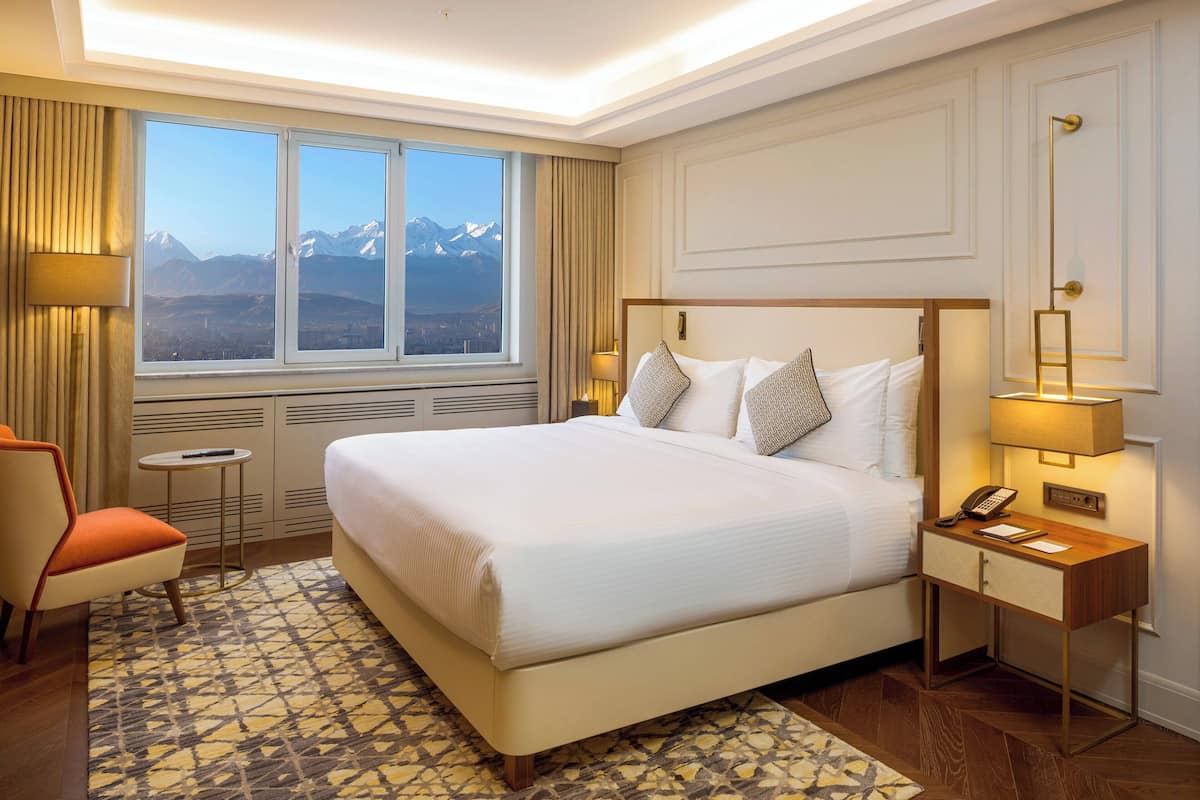 A hotel room with a large bed, a bedside table with a lamp, a chair, and a view of mountains through the window.