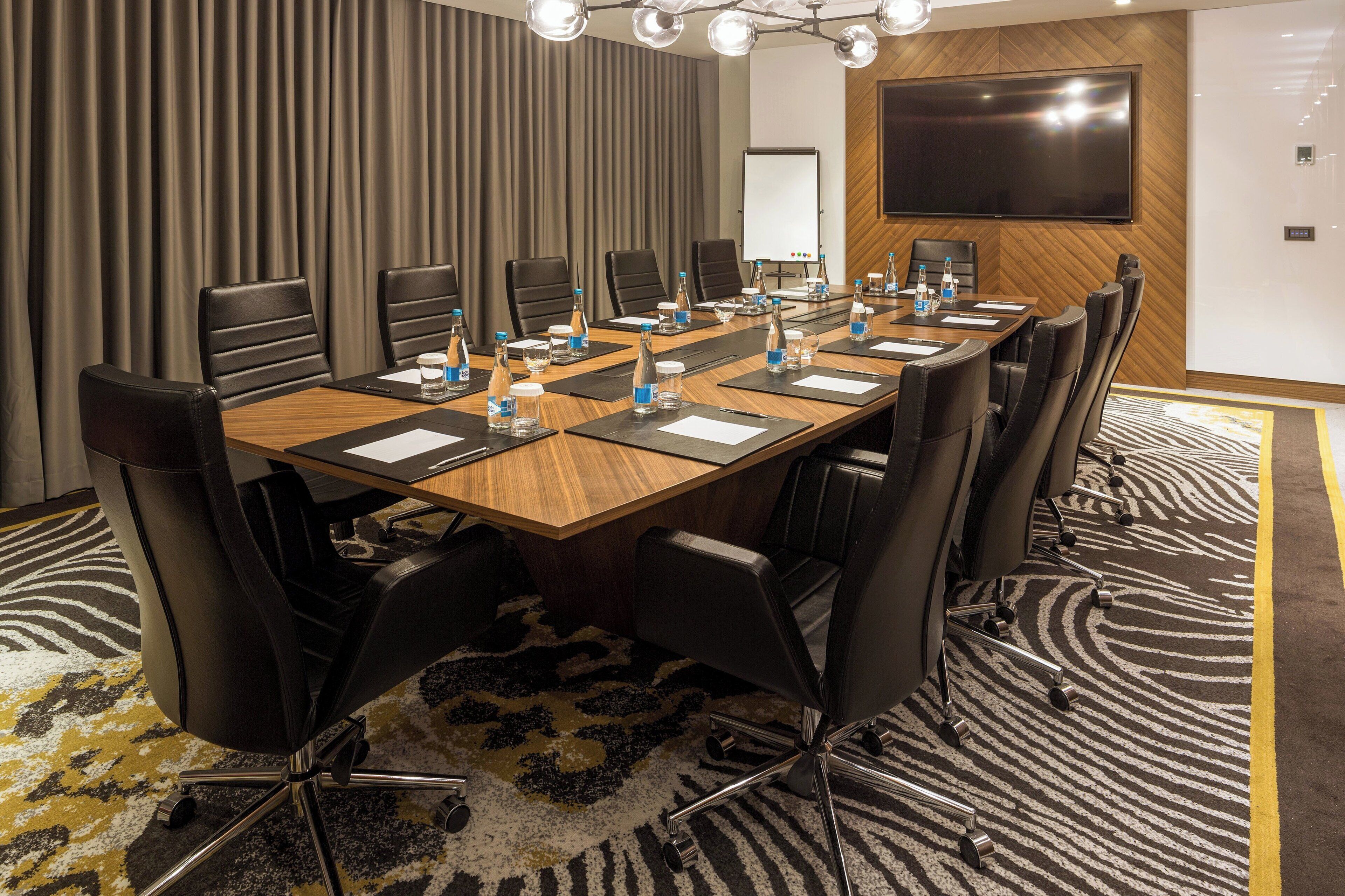 A conference room with a long table, multiple chairs, water bottles, and a large screen on the wall.