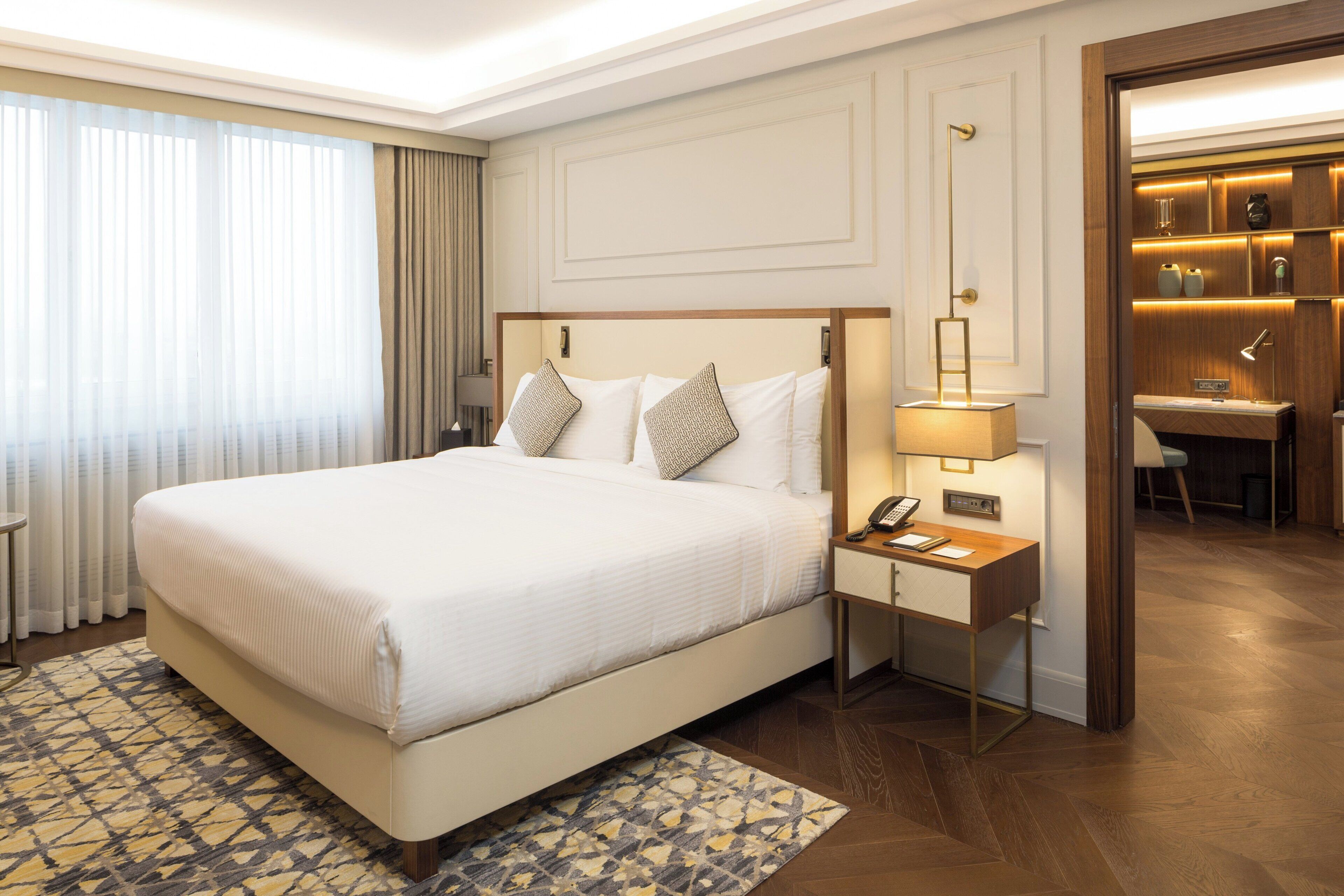 Una habitación de hotel con una cama grande, una mesita de noche con lámpara, un escritorio y un espejo.