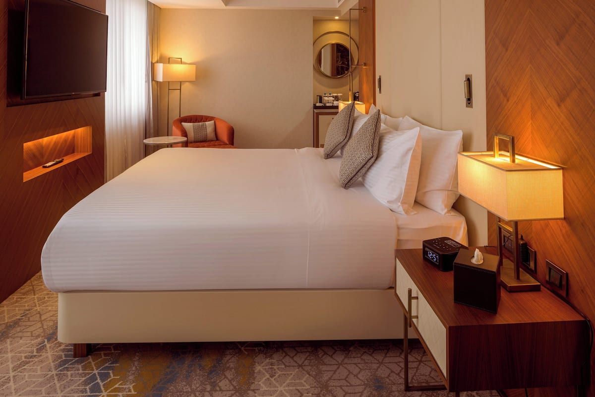 A hotel room with a large bed, bedside table with a lamp, a TV mounted on the wall, and a small seating area with a chair and table.