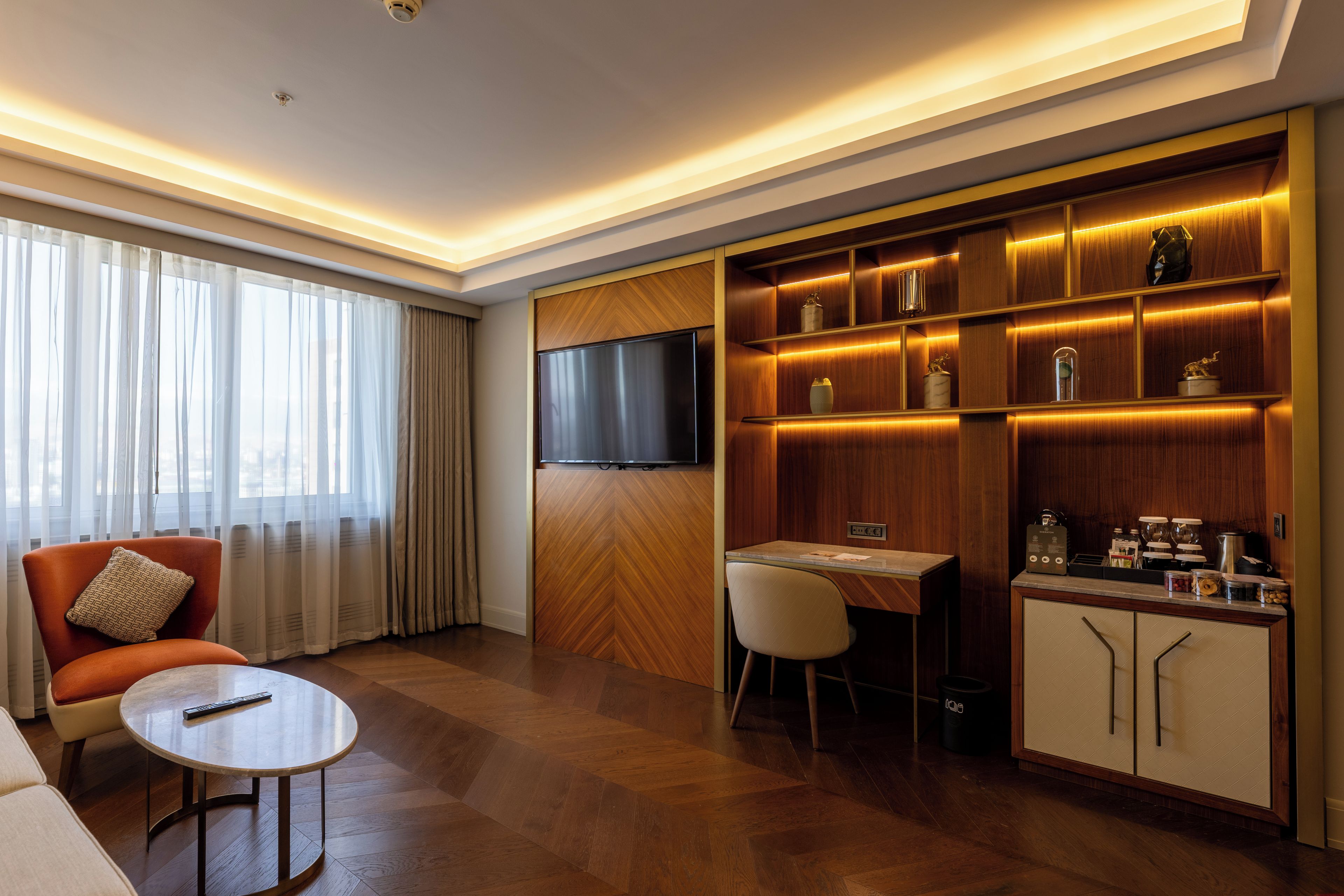 Ein modernes Hotelzimmer mit einer Couch, einem runden Tisch, einem an der Wand befestigten Fernseher und einer eingebauten Minibar.