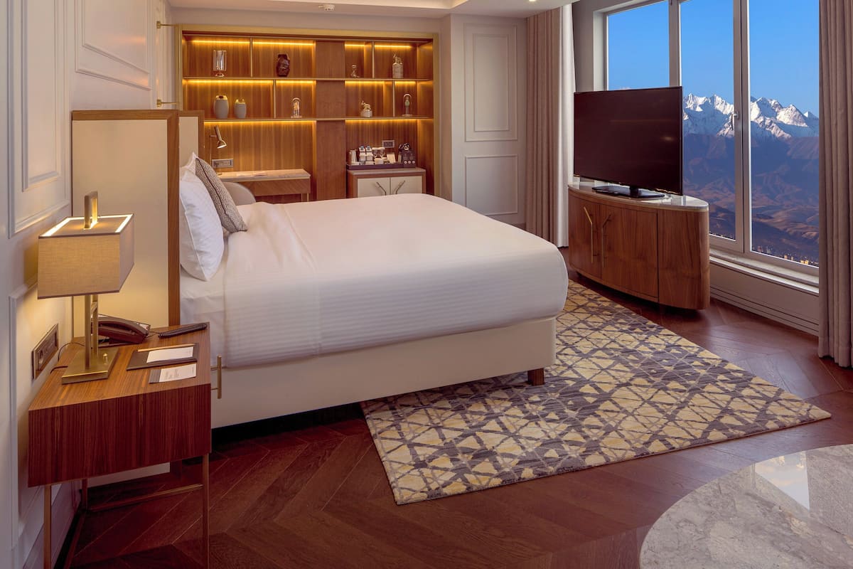A hotel room with a large bed, a TV, a desk with a lamp, a bedside table, a patterned rug, and a view of mountains through the window.
