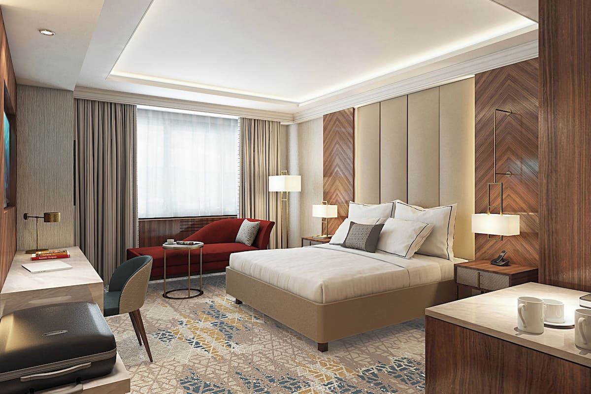 A hotel room with a bed, a desk, a chair, a red sofa, and a small table with a lamp.