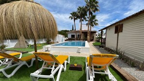A poolside area with artificial grass, yellow and white lounge chairs, a thatched umbrella, and a house in the background.