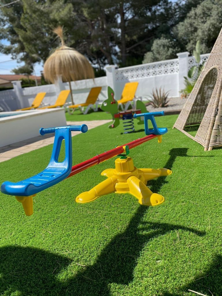 Un espace de jeux pour enfants avec une balançoire, un ensemble de jeux à bascule et une petite structure d’escalade sur une pelouse artificielle.