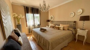 A bedroom with a large bed, a bedside table, a nightstand, a bench, a chandelier, and a window with curtains.
