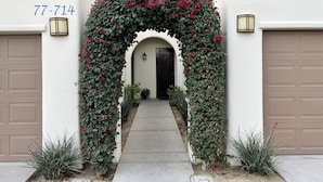 A white building with a beige garage door, a red flower archway, and a number '77-714' on the wall. - Luxurious Ground floor Villa with 2 Master Bedrms and views (La Quinta)