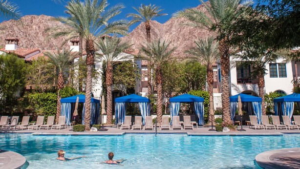 A pool area with lounge chairs and umbrellas, surrounded by palm trees and buildings with a mountainous backdrop. - Luxurious Ground floor Villa with 2 Master Bedrms and views (La Quinta)