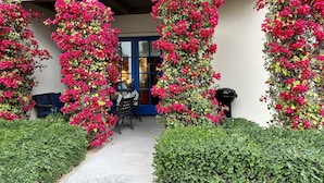A building facade with vibrant pink bougainvillea climbing up the walls, a blue door, and a small outdoor seating area with two chairs. - Luxurious Ground floor Villa with 2 Master Bedrms and views (La Quinta)