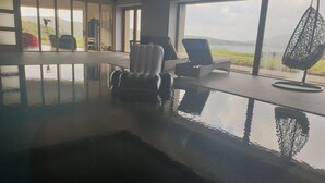 An indoor swimming pool with lounge chairs and a hanging chair, large windows with a view of the outdoors. - Fab View (Dingle)