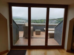 A room with a large sliding glass door leading to an outdoor balcony with wicker furniture and a view of a town and hills.