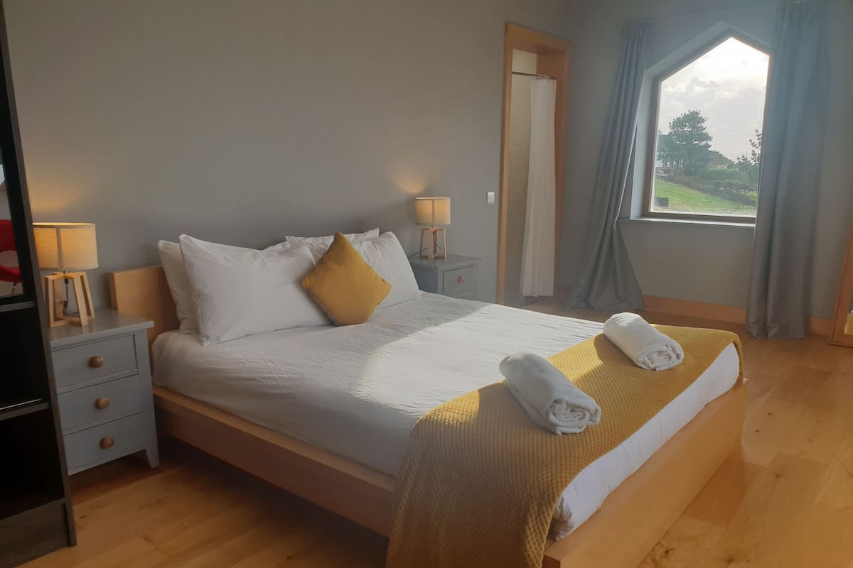 Cama bem arrumada com roupa de cama branca e amarela, cabeceira de madeira e uma janela com cortinas.