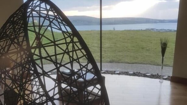 A hanging chair with a geometric design in a room with a large window, wooden flooring, and a view of a grassy field and water. - Fab View (Dingle)