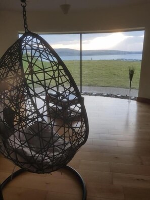 A hanging chair with a geometric design in a room with a large window, wooden flooring, and a view of a grassy field and water. - Fab View (Dingle)