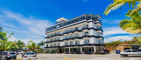 A multi-story hotel with a modern design, featuring balconies on each floor, a parking lot with several cars, and palm trees in the vicinity.