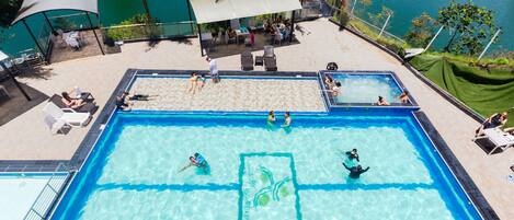 A rectangular swimming pool with clear blue water, surrounded by lounge chairs and umbrellas, with a view of a lake and palm trees.