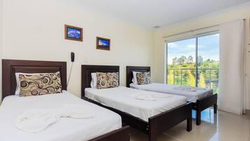 A hotel room with three beds, each with a wooden headboard and patterned pillows. There are two framed pictures on the wall, and a balcony with a view of trees and a clear sky.