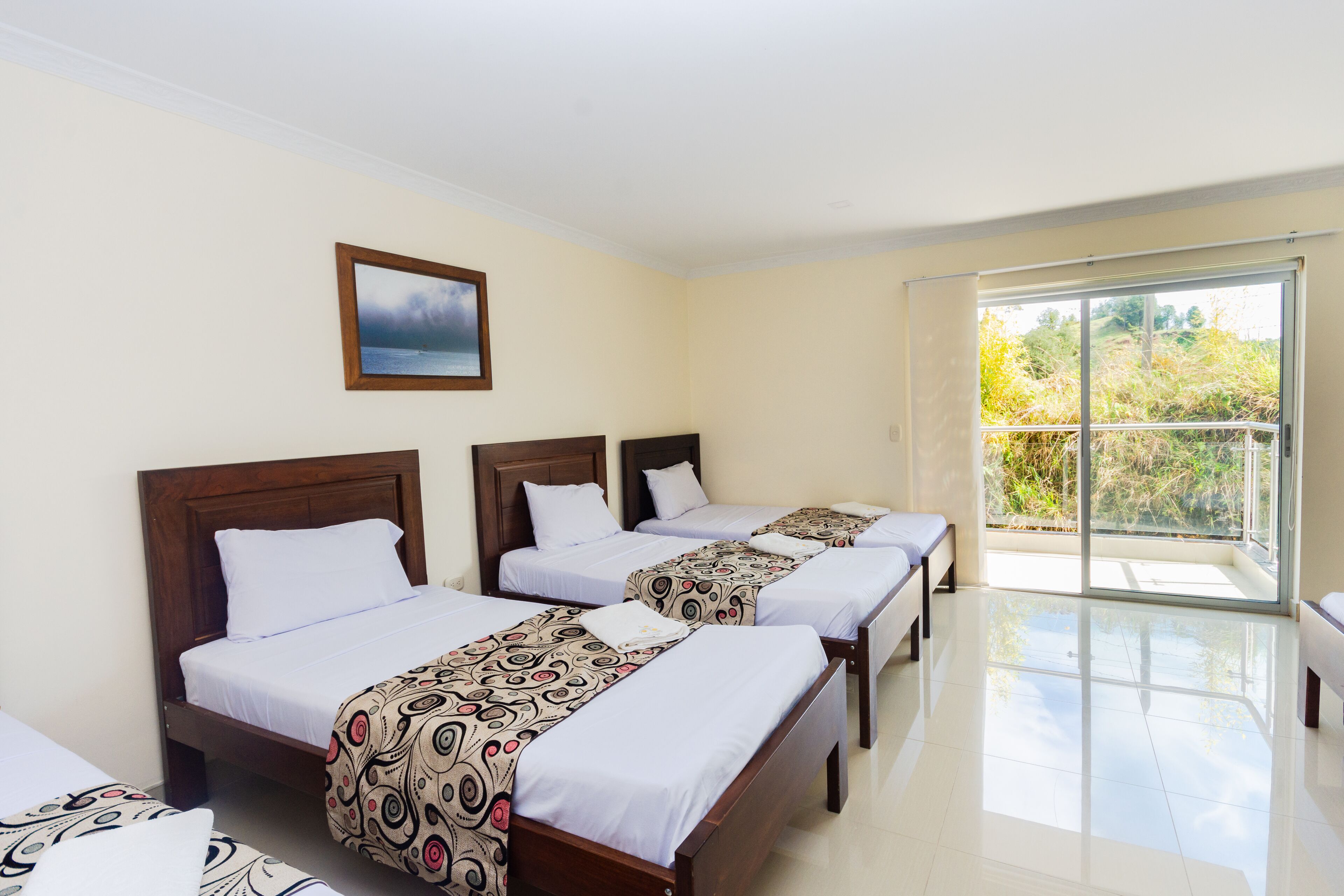 A hotel room with three beds, a balcony with a view of trees, and a painting on the wall.