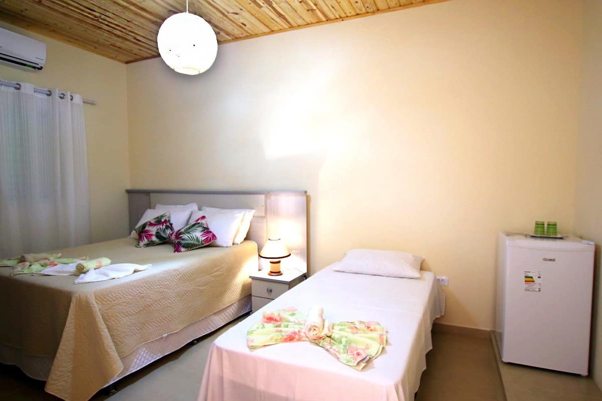 A hotel room with two beds, a nightstand with a lamp, a refrigerator, and an air conditioning unit.