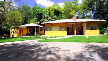 Um edifício amarelo com telhado de telhas, varanda coberta e um pequeno jardim.