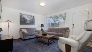 A living room with a grey sofa, a wooden coffee table, a white armchair, and a large window with a view of palm trees. - Convenient 2 bedroom, walk to Bart, 10 min to Berkeley (Richmond)