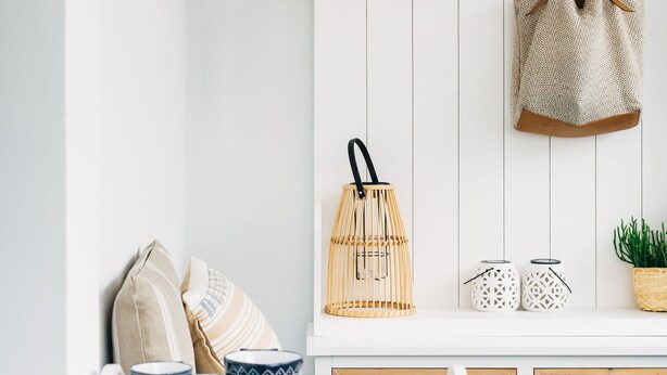 Pared blanca con un estante de madera que sostiene un sombrero, una cesta tejida y un cuadro enmarco. Debajo, una mesa de mimbre blanca sostiene una bandeja con objetos decorativos.