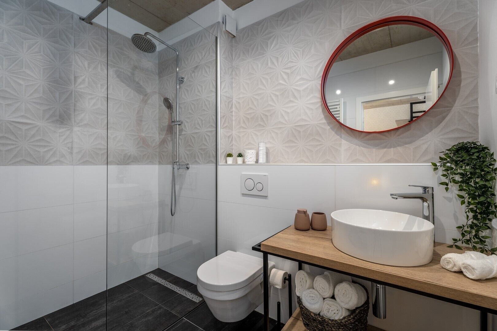 Un baño moderno con ducha envuelta en vidrio, inodoro blanco, lavamanos redondo con encimera de madera y espejo con marco rojo.