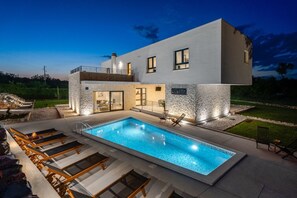 Une maison moderne à deux étages, dotée d’une piscine, d’un espace salon extérieur et d’une façade en pierre.