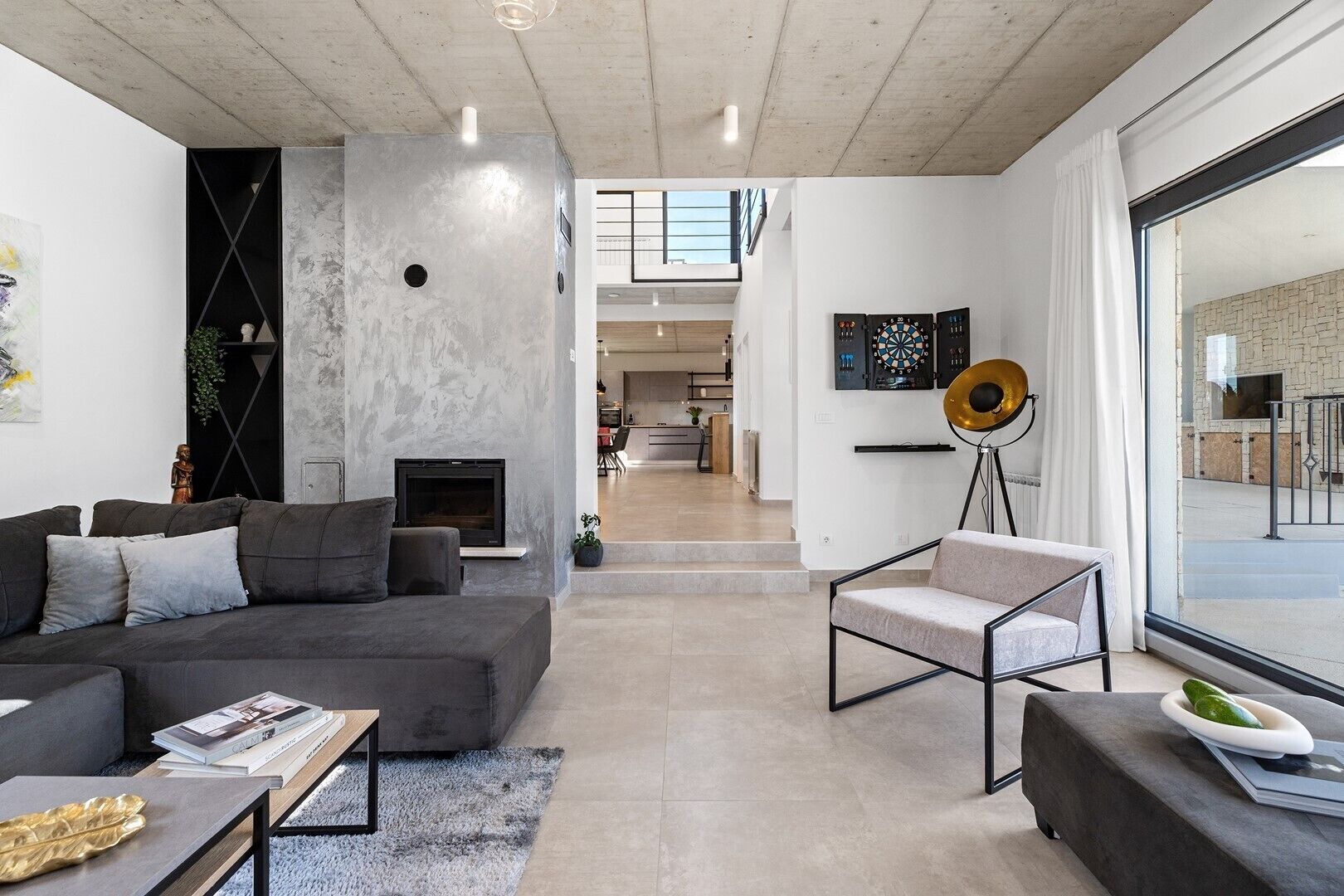A modern living room with a grey sofa, a small table, and a floor lamp. There is a fireplace and a view of the kitchen in the background.