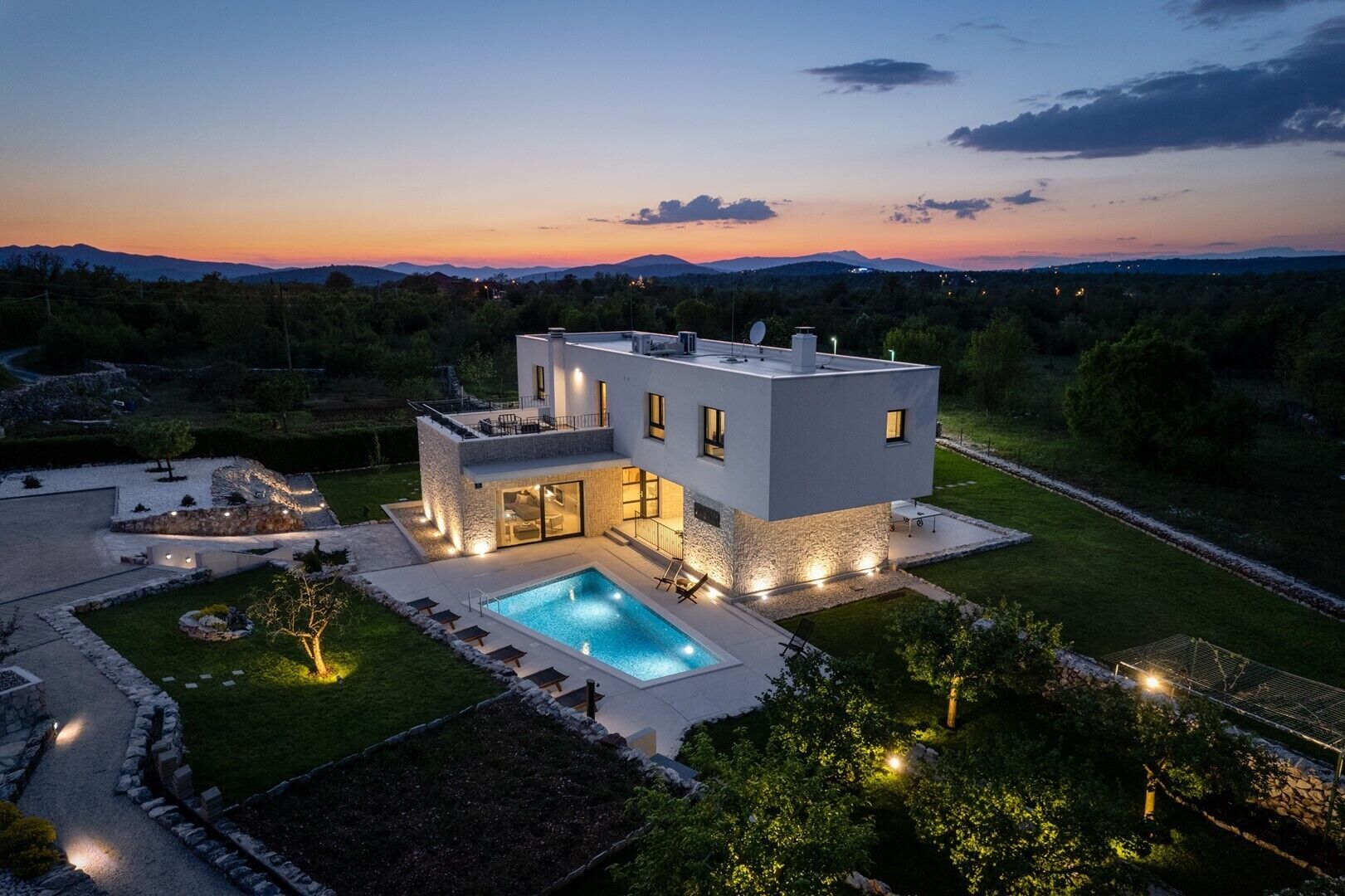 A modern two-story house with a swimming pool, surrounded by a well-maintained garden and a view of distant mountains.
