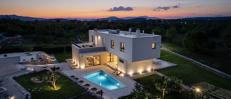 A modern two-story house with a swimming pool, surrounded by a well-maintained garden and a view of distant mountains.