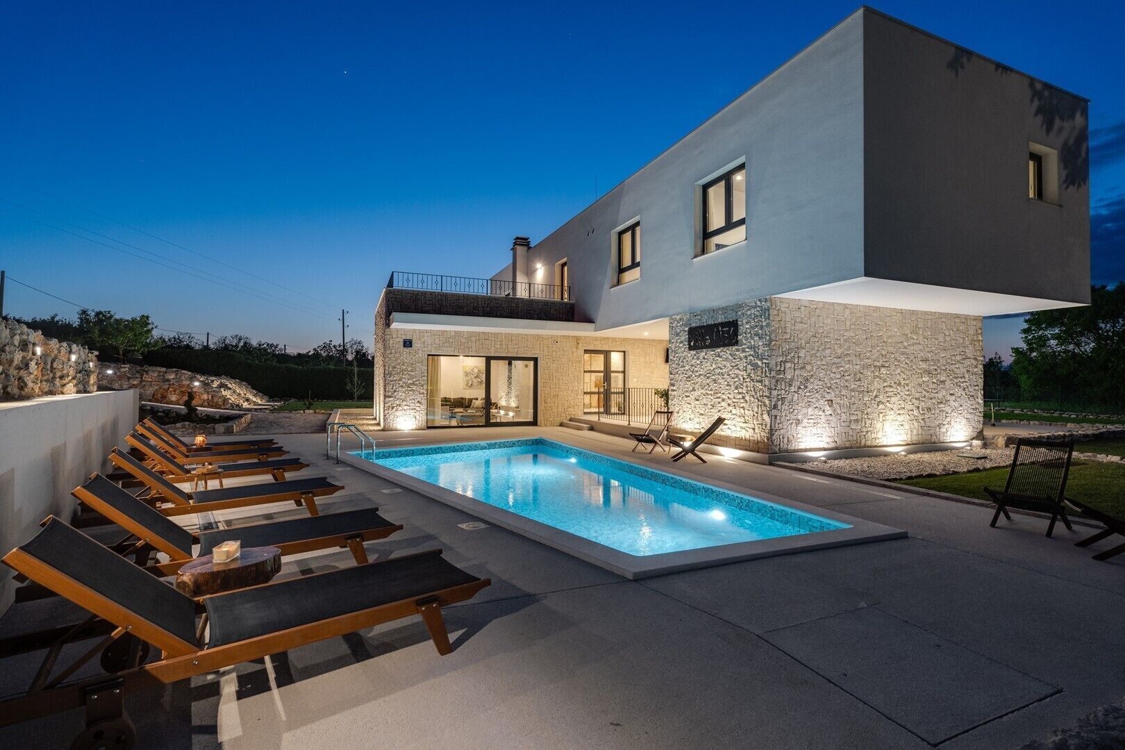 A modern two-story house with a swimming pool, lounge chairs, and stone walls.