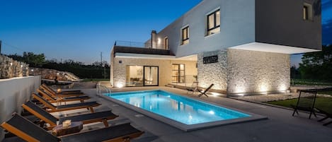 A modern two-story house with a swimming pool, lounge chairs, and stone walls.