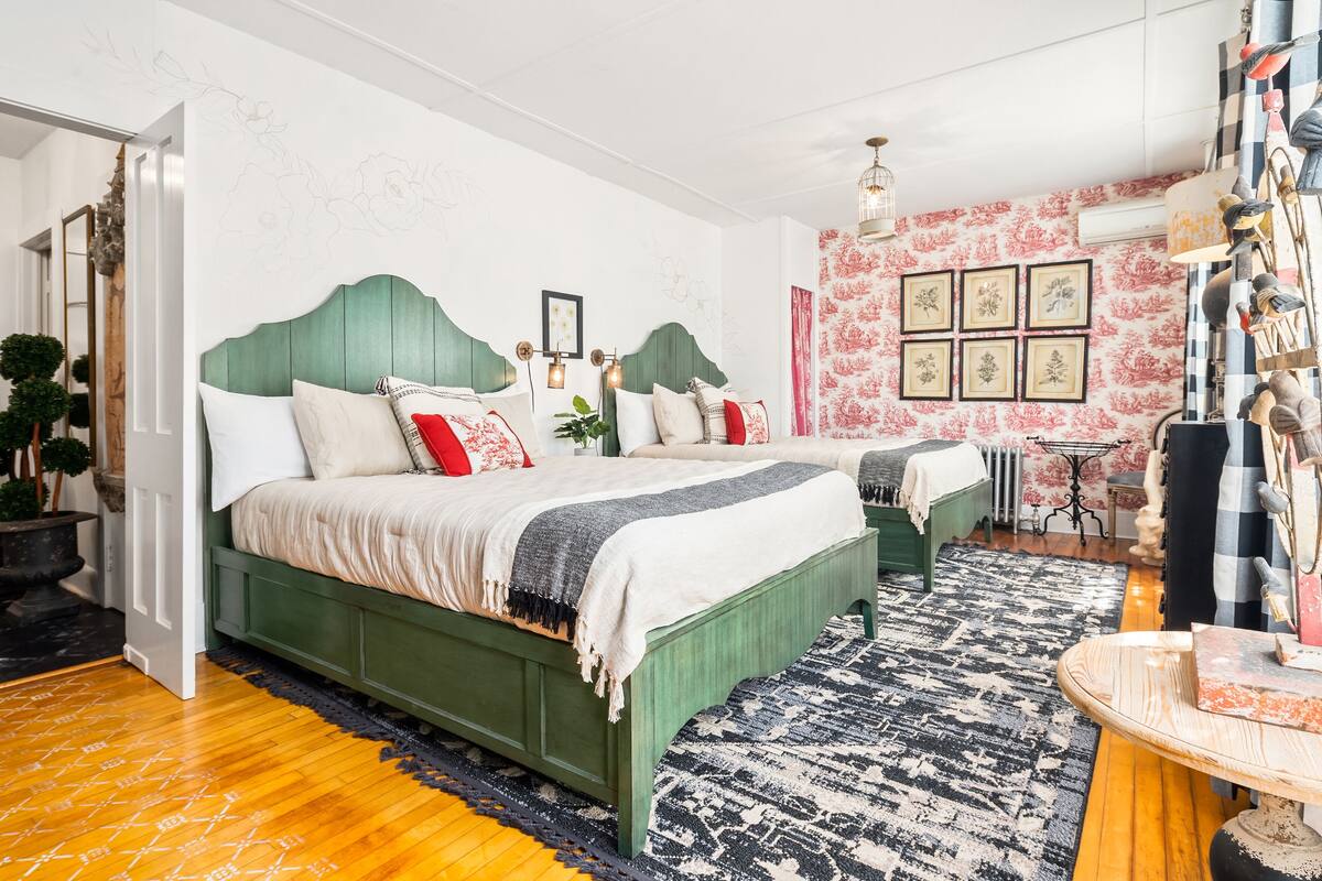 Ein Schlafzimmer mit zwei mit grünem Stoff gepolsterten Betten, einem gemusterten Teppich und einer Wand mit gerahmten Kunstwerken.
