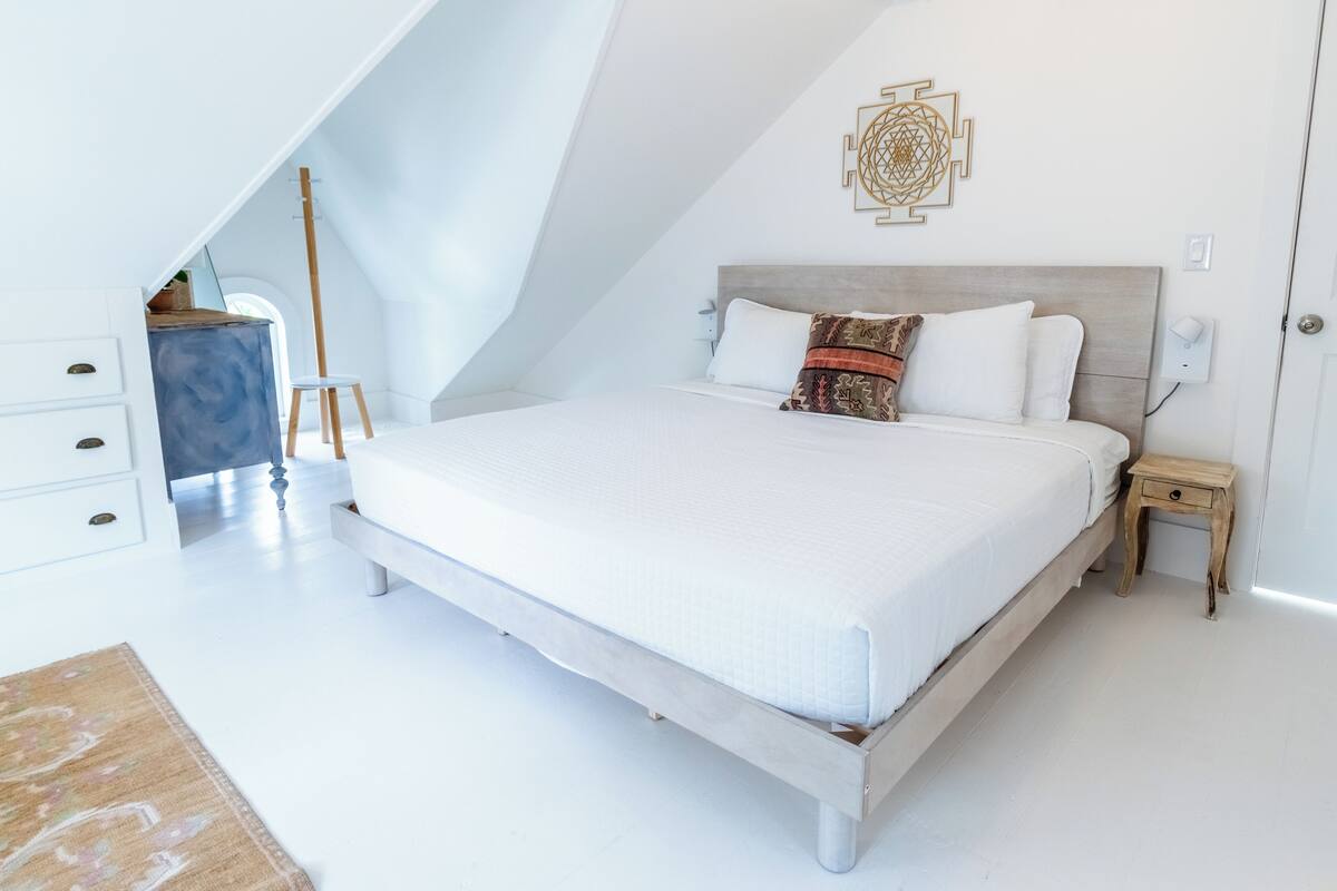 Ein Schlafzimmer mit schräg gedachter Decke, einem Bett mit weißer Bettwäsche, einem Nachttisch aus Holz und einem Wandbild.