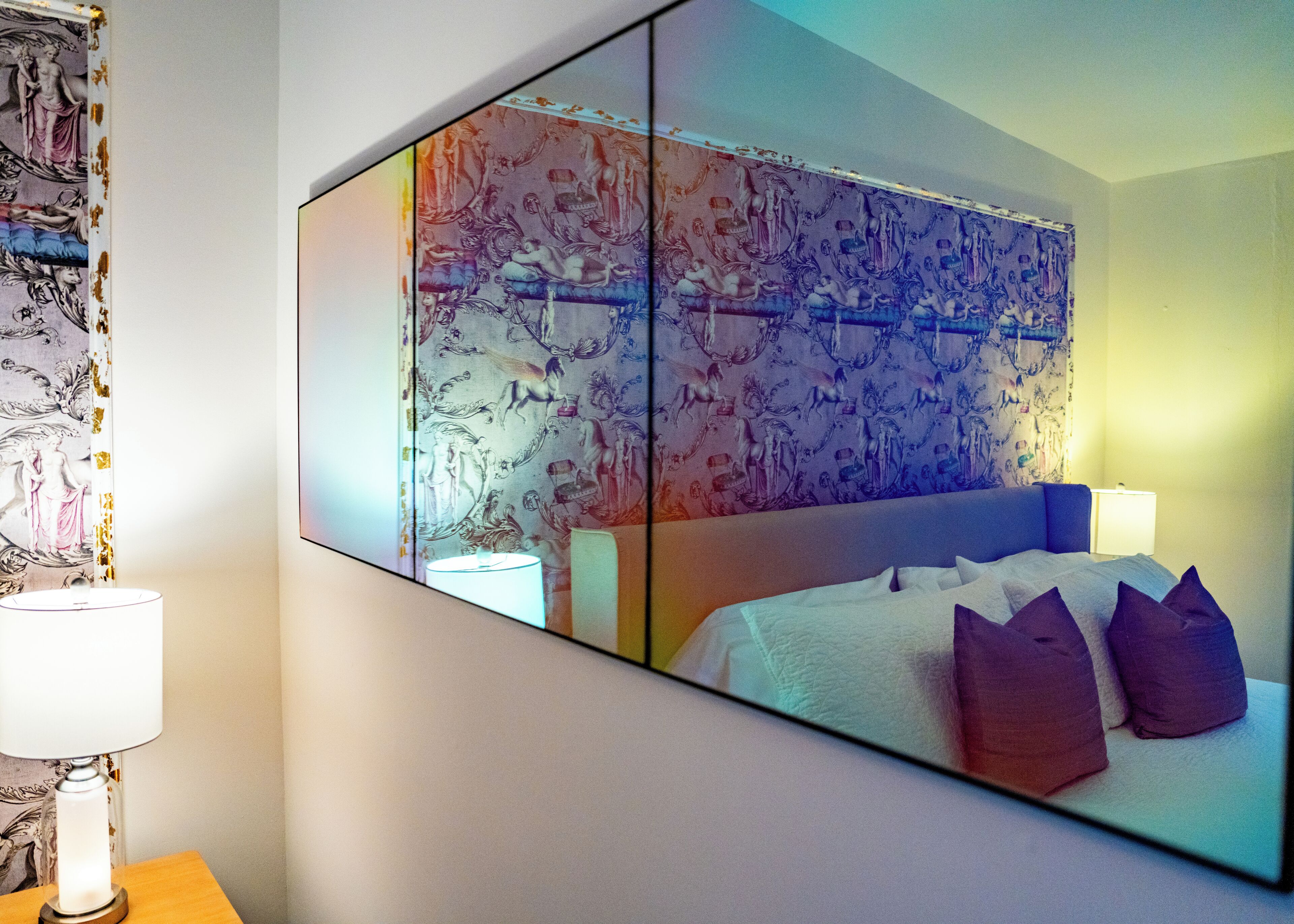 Ein Hotelzimmer mit einem großen Bett, einem Nachttisch mit Lampe und einem an der Wand befestigten Spiegel, der den Raum widerspiegelt.