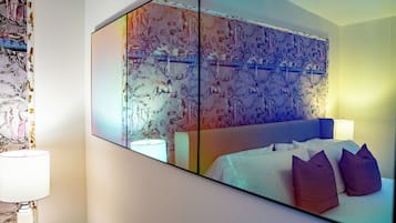 A hotel room with a large bed, a bedside table with a lamp, and a wall-mounted mirror reflecting the room.