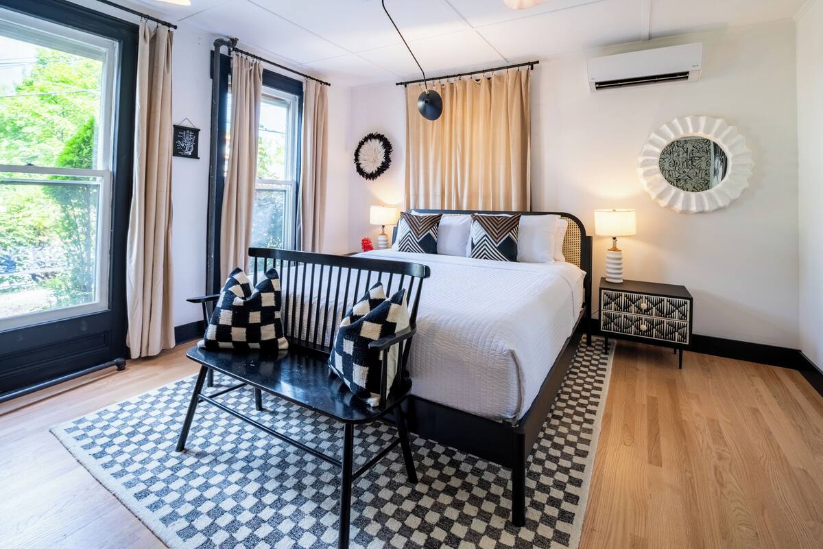Ein modernes Schlafzimmer mit einem großen Bett, einem Nachttisch mit Lampe, Blick auf Grünfläche und einem karierten Teppich.