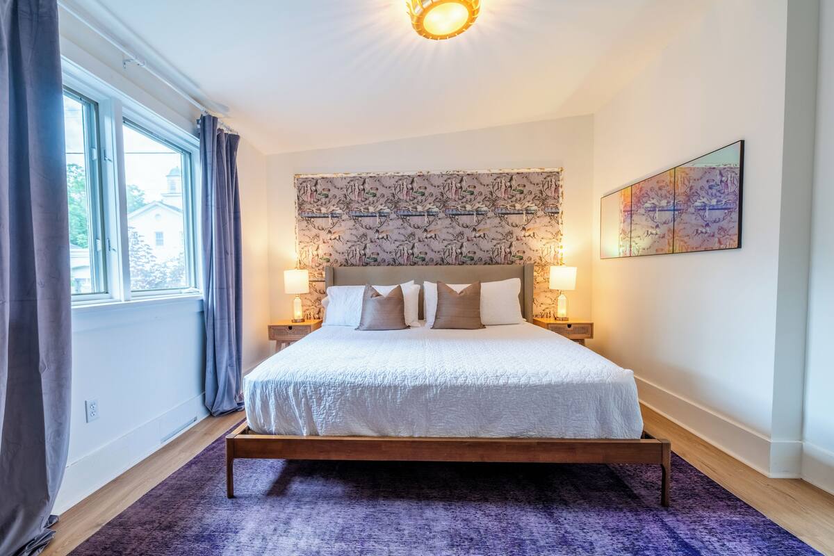 Ein Schlafzimmer mit einem Bett, zwei Nachttischlampen, einem gemusterten Kopfteil, einem Fenster mit Vorhängen und einem Wandbild.