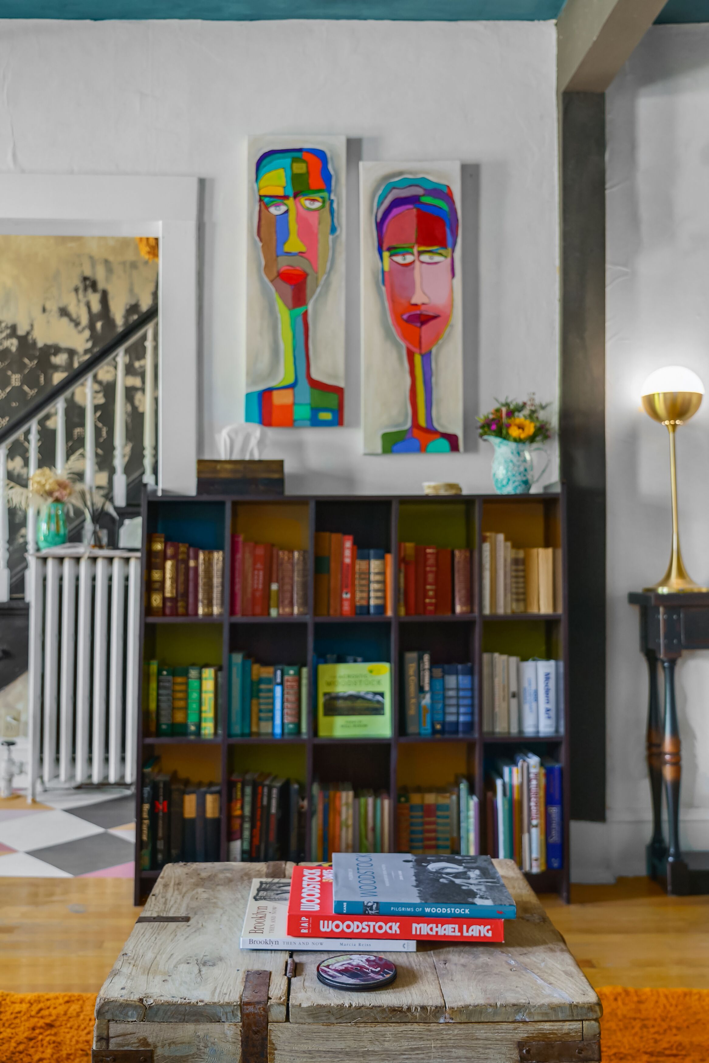 A room with a wooden coffee table, a bookshelf filled with books, and two colorful abstract paintings on the wall.