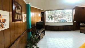 A room with a wooden interior, a large window, a yellow sofa, and a TV on a wooden stand. - Hostal Casa Lantana Usaquén - Hostel (Bogotá)