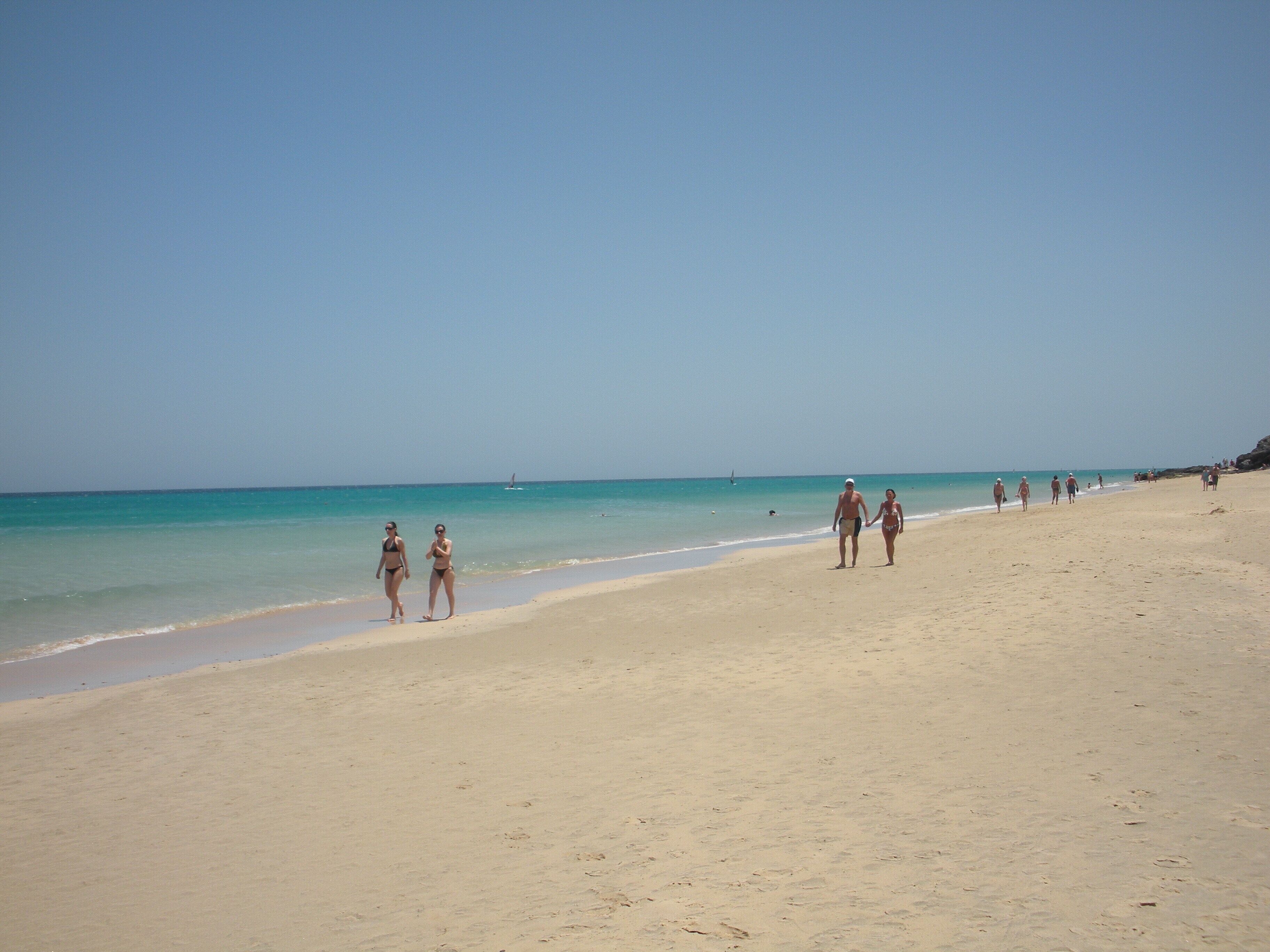 Una amplia playa de arena con agua turquesa cristalina y algunas personas caminando por la orilla.