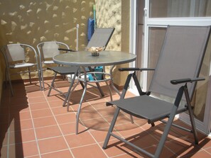 A patio with a round table and four chairs, a blue trash bin, and a window with a screen. - Beachfront studio apartment (Costa Calma)