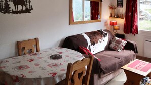 A cozy living room with a brown sofa, a wooden table with a red tablecloth, and a window with red curtains. - Lovely 2-room apartment, slopeside, sleeps 6 (Les Orres)