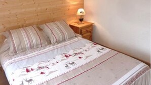 A bedroom with a wooden wall, a bed with a patterned bedspread, a nightstand with a lamp, and a small picture frame on the wall. - Lovely 2-room apartment, slopeside, sleeps 6 (Les Orres)