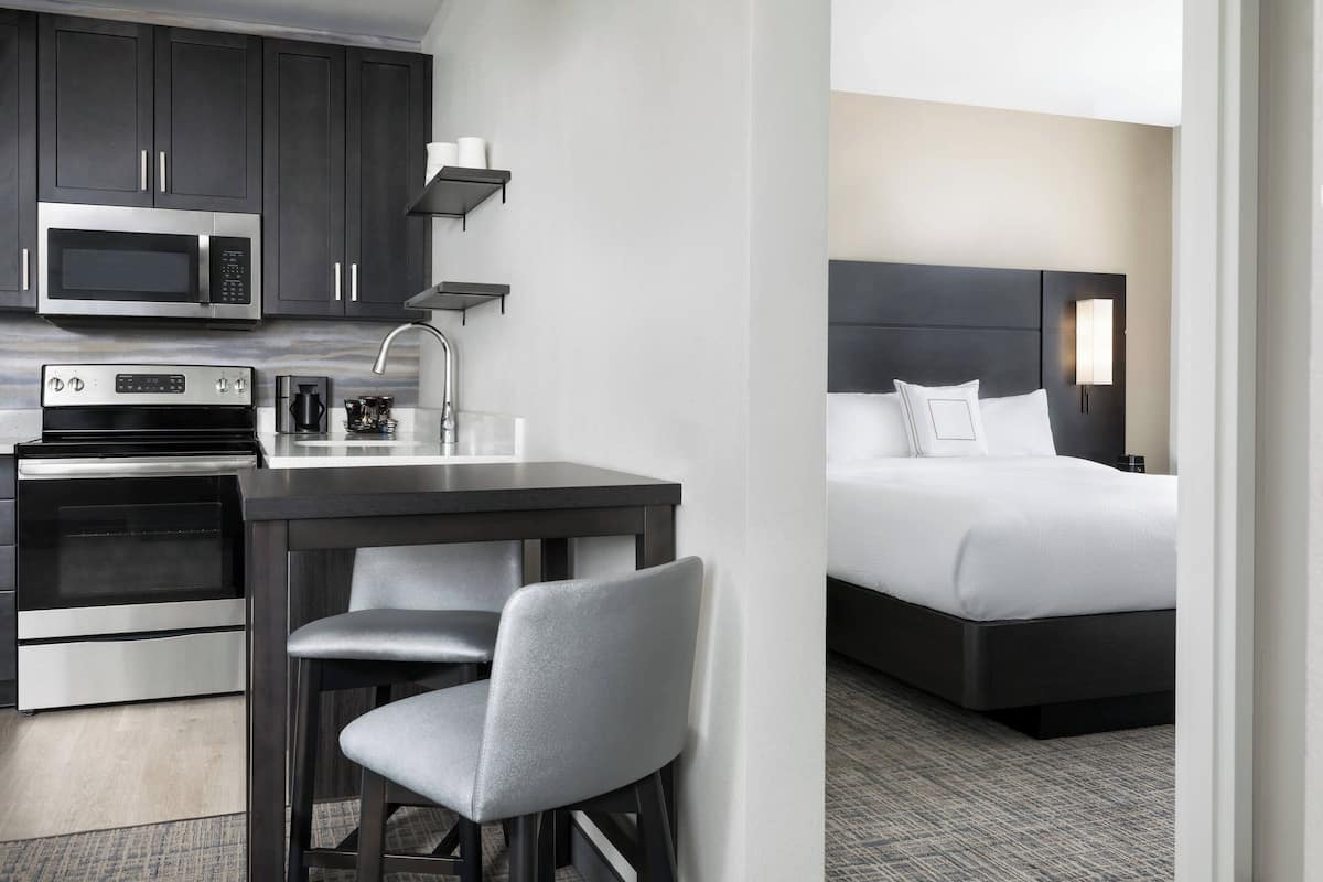 Ein modernes Hotelzimmer mit Küchenzeile, einem Bett mit dunklem Kopfteil und einer kleinen Essecke mit Barhocker und Stuhl.