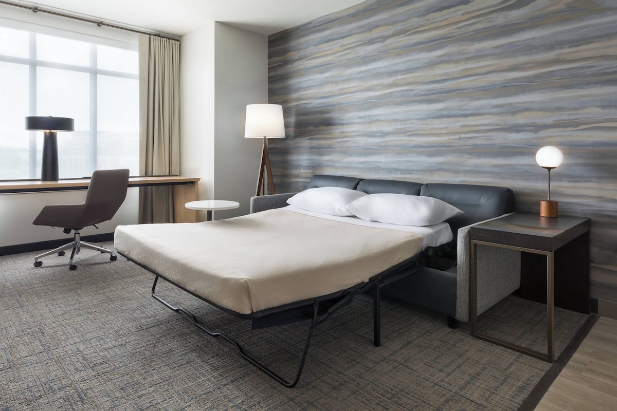 Ein Hotelzimmer mit Bett, Schreibtisch, Stuhl, Lampe und Blick auf die Stadt durch das Fenster.